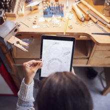 Overhead View Of Female Jeweller Comparing Ring With Drawn Design On Digital Tablet In Studio